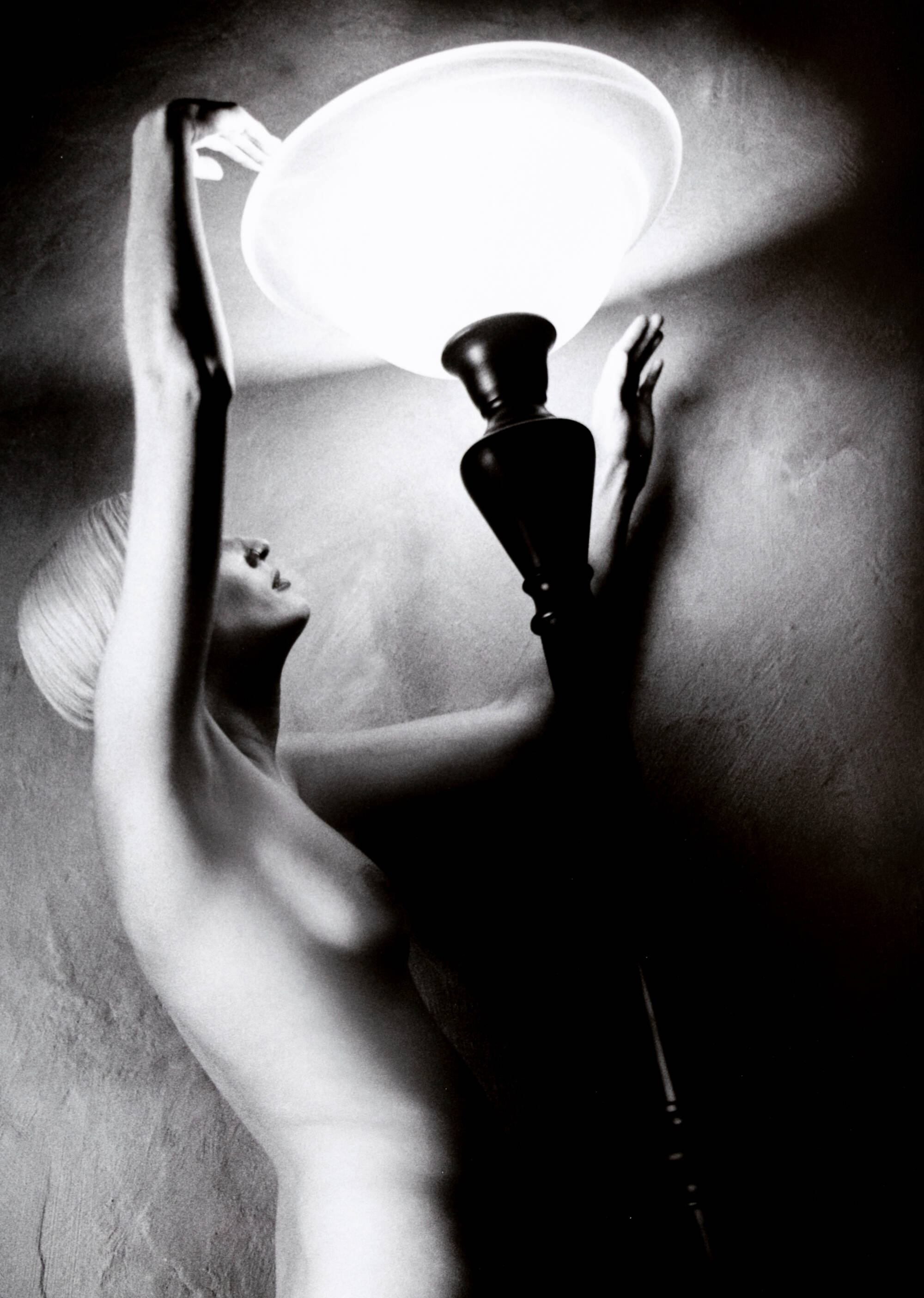 Black and white photograph of a nude woman touching the top of a floor lamp.